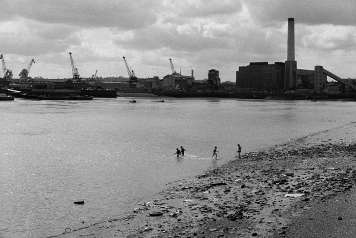 Black and white photograph of a run down waterfront of the Isle of Dogs in the 1980s. Many derelict industrial buildings and a few cranes are visible.